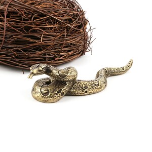 Op de afbeelding: Een gedetailleerd messing slangenbeeldje met een getextureerd lichaam en een gespleten tong. De slang is opgerold en ligt op een wit oppervlak. Een nest van verweven bruine twijgen bevindt zich op de achtergrond.