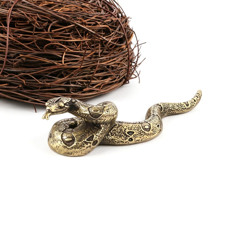 Op de afbeelding: Een gedetailleerd messing slangenbeeldje met een getextureerd lichaam en een gespleten tong. De slang is opgerold en ligt op een wit oppervlak. Een nest van verweven bruine twijgen bevindt zich op de achtergrond.