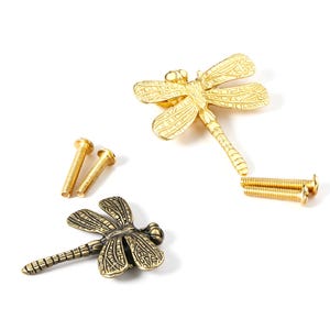 May include: Two dragonfly-shaped metal cabinet knobs, one in a bright gold finish and one in an antique brass finish, each featuring detailed wing textures and a segmented body, accompanied by four matching threaded metal mounting screws.