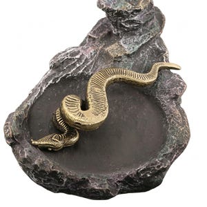 Op de afbeelding: Een decoratieve asbak met een bronzen slang die zich in een donker, getextureerd bekken kronkelt. Het bekken is geplaatst in een rotsachtige structuur met groene en bruine tinten. De schubben van de slang zijn gedetailleerd.