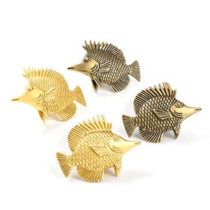 May include: Four metal fish-shaped decorative objects arranged in a group. Two of the fish have a bright, polished gold finish, while the other two feature a darker, antique brass finish. Each fish displays detailed scale patterns and fins.