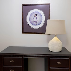 May include: A dark wood desk with multiple drawers, a table lamp with a textured white base and a beige drum shade, and a square framed print featuring a circular illustration of a figure in a purple outfit against a light purple background.