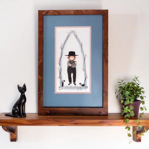 May include: A framed illustration featuring a person in a black hat and overalls standing within a pointed archway decorated with pink flowers, flanked by two small black cats, all enclosed in a dark wood frame with a blue and pink mat.