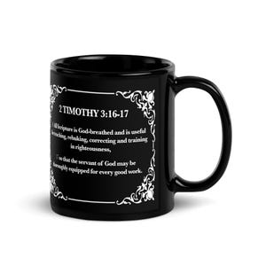 May include: A black ceramic mug with a white decorative border and text. The text reads "2 TIMOTHY 3:16-17" and a passage from the Bible. The mug has a large handle and is designed for hot beverages.