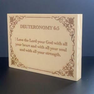 May include: A rectangular wooden block with the text "DEUTERONOMY 6:5" and a Bible verse engraved in a decorative border. The block is light-colored with a natural wood grain.