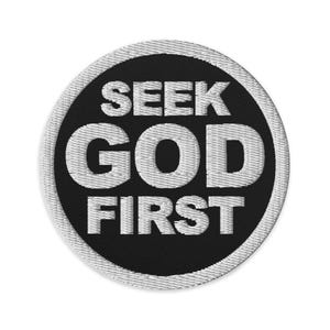May include: A round, embroidered patch with the words "SEEK GOD FIRST" in white, block letters on a black background. The patch has a white border and is likely designed to be sewn onto clothing or accessories.