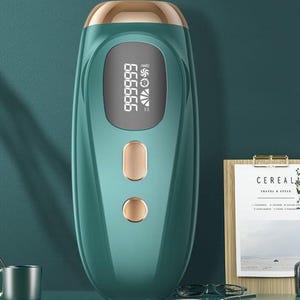 May include: A teal and gold handheld hair removal device with a digital display showing a six-digit number. The device has two gold-colored buttons and a gold-colored top. The background is a dark teal color.