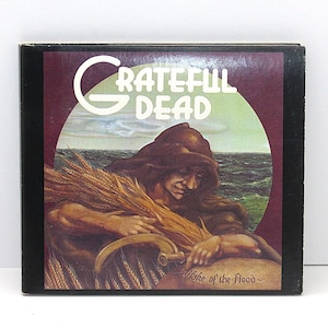 May include: A square album cover featuring the band name "Grateful Dead" in white lettering. The artwork depicts a person holding wheat, with a sea and sky background. The album title "Wake of the Flood" is also visible.