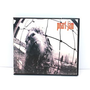 May include: A CD case featuring a black and white image of a sheep behind a fence. The band name "pearl jam" is printed in orange at the top right corner. The image has a vintage, grunge aesthetic.