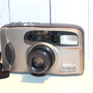 Pode incluir: Câmera Nikon One-Touch Zoom 70 AF prateada e preta com lente 38-70mm, alça preta e rolo de filme Kodak Max 400. A câmera possui flash embutido e foi projetada para facilitar o uso.