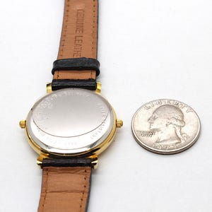 May include: A gold-toned wristwatch with a black leather band and a brown leather strap. The watch face is not visible. A quarter coin is next to the watch. The watch is marked "Genuine Leather" on the strap.