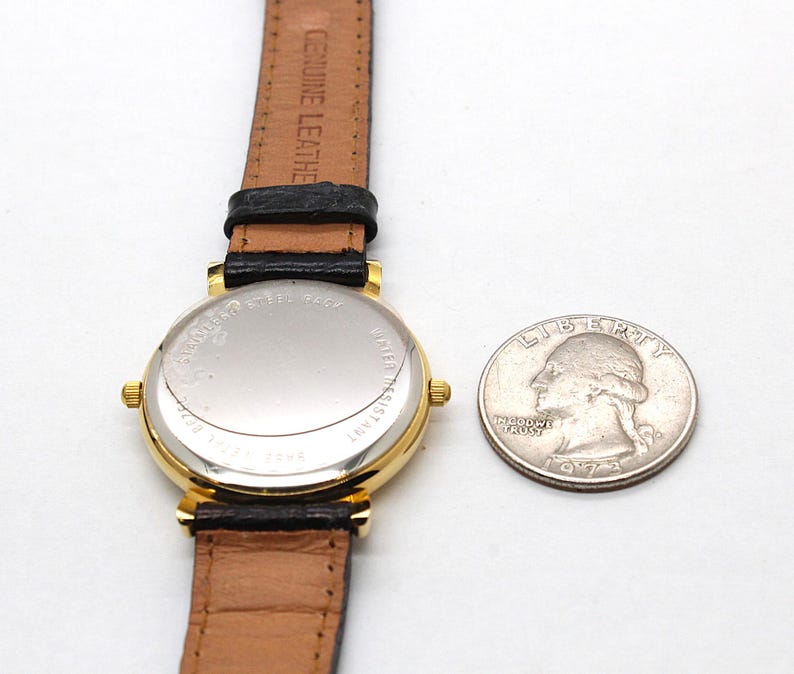 May include: A gold-toned wristwatch with a black leather band and a brown leather strap. The watch face is not visible. A quarter coin is next to the watch. The watch is marked "Genuine Leather" on the strap.