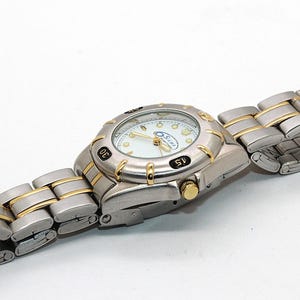 May include: A silver-toned wristwatch with gold accents. The watch face is white with black numerals and gold hands. The watch band is silver with gold-colored links. The watch is on a white surface.