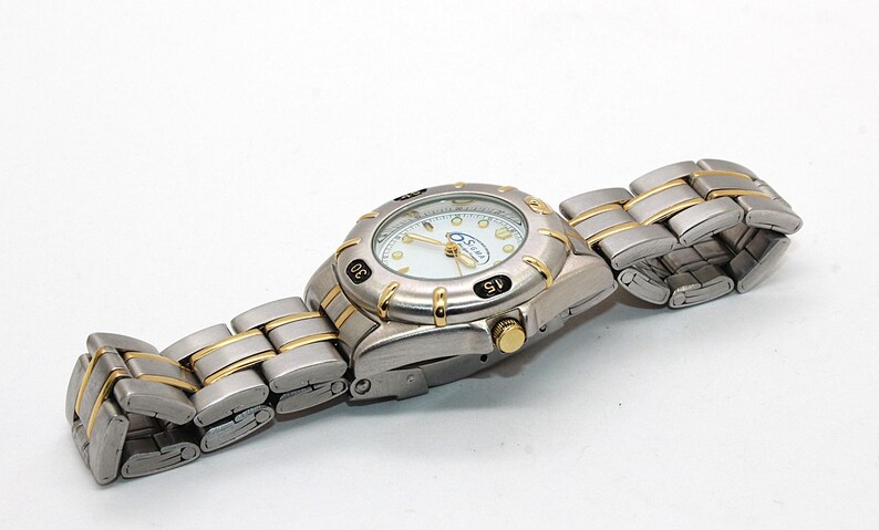 May include: A silver-toned wristwatch with gold accents. The watch face is white with black numerals and gold hands. The watch band is silver with gold-colored links. The watch is on a white surface.