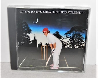 May include: A CD case with a black spine and a clear front cover. The front cover features a photo of Elton John in a red hat and white clothing, playing cricket on a green field. The text "ELTON JOHN'S GREATEST HITS VOLUME II" is printed in white on a black background at the top of the cover.