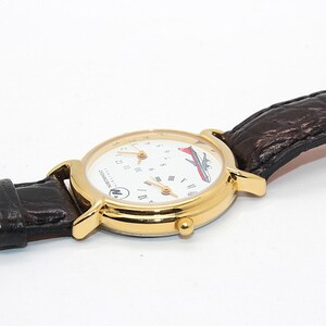 May include: A gold-toned wristwatch with a white face, featuring a red and white airplane graphic. The watch has a brown leather band and a round face with multiple dials. The watch is set against a white background.