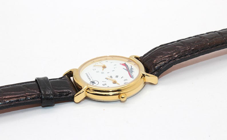 May include: A gold-toned wristwatch with a white face, featuring a red and white airplane graphic. The watch has a brown leather band and a round face with multiple dials. The watch is set against a white background.