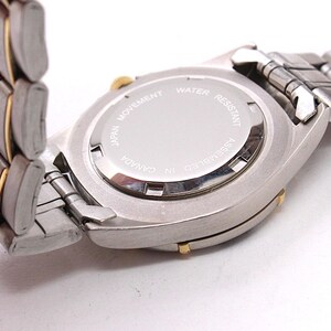 May include: Close-up of a silver and gold-toned wristwatch. The watch has a silver band with gold accents. The watch face is silver with the text "JAPAN MOVEMENT WATER RESISTANT ASSEMBLED IN CANADA".
