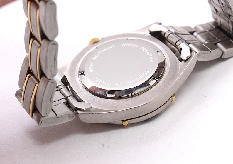 May include: Close-up of a silver and gold-toned wristwatch. The watch has a silver band with gold accents. The watch face is silver with the text "JAPAN MOVEMENT WATER RESISTANT ASSEMBLED IN CANADA".