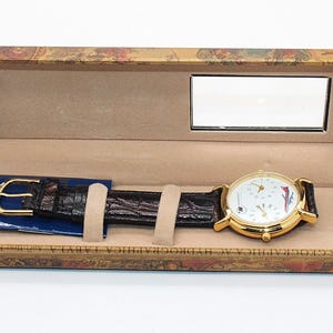 May include: A gold-toned wristwatch with a white face and black leather band rests in an open decorative box. The watch face features multiple dials and red and blue accents. The box has a mirror and a patterned exterior.