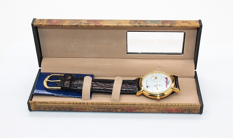 May include: A gold-toned wristwatch with a white face and black leather band rests in an open decorative box. The watch face features multiple dials and red and blue accents. The box has a mirror and a patterned exterior.