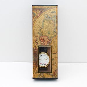 May include: A wristwatch with a black leather band and a white face featuring a red and white airplane design. The watch is displayed in a rectangular box decorated with a vintage world map.