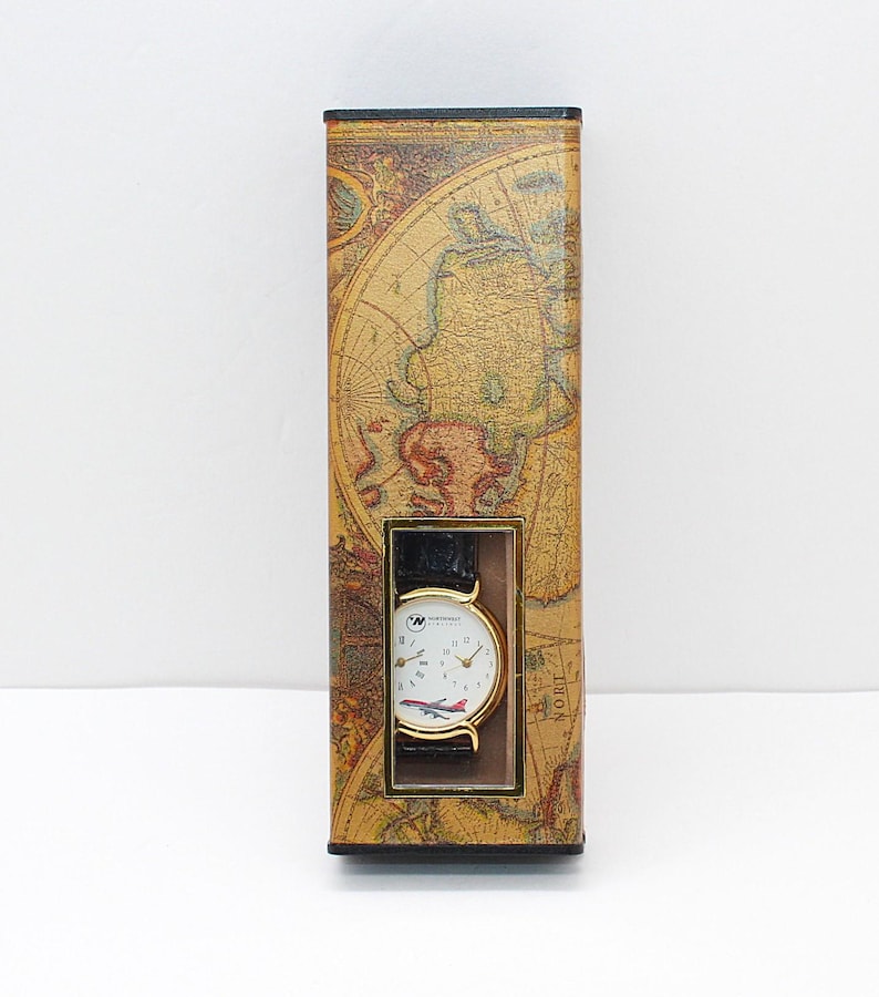May include: A wristwatch with a black leather band and a white face featuring a red and white airplane design. The watch is displayed in a rectangular box decorated with a vintage world map.