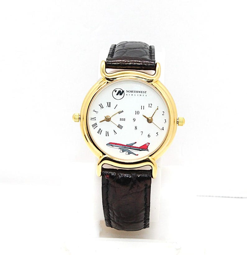 May include: A gold-toned wristwatch with a white face featuring Roman numerals and a small airplane graphic. The watch has a brown leather band and the Northwest Airlines logo. The watch has two separate dials.
