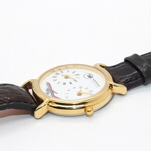 May include: A gold-toned wristwatch with a white face featuring Roman numerals and a red and white airplane graphic. The watch has a brown leather band. The watch face also has the text "NORTHWEST AIRLINES".