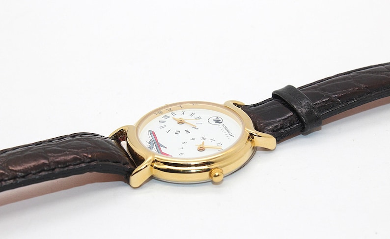 May include: A gold-toned wristwatch with a white face featuring Roman numerals and a red and white airplane graphic. The watch has a brown leather band. The watch face also has the text "NORTHWEST AIRLINES".