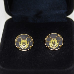 May include: A pair of gold Mickey Mouse cufflinks in a black velvet box. The cufflinks are round and feature a black background with a gold Mickey Mouse head.