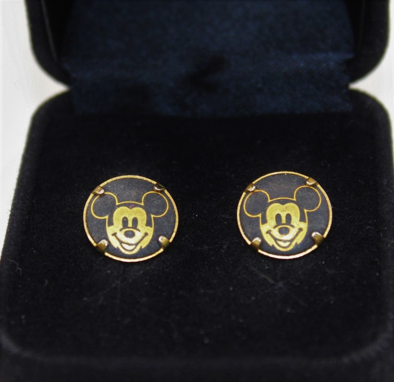 May include: A pair of gold Mickey Mouse cufflinks in a black velvet box. The cufflinks are round and feature a black background with a gold Mickey Mouse head.