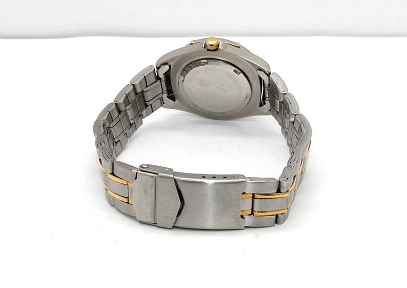 May include: A stainless steel wristwatch with a silver and gold-tone band. The watch has a round face and a clasp closure. The back of the watch is visible, showing the battery compartment and other details. The watch band is approximately 8 inches long.