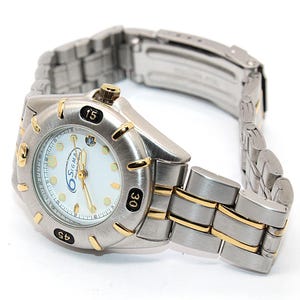May include: A silver-toned wristwatch with gold accents. The watch face is white with black numerals and the brand name "Sigma". The watch band is silver with gold-toned links. The watch is angled to show the face and band.