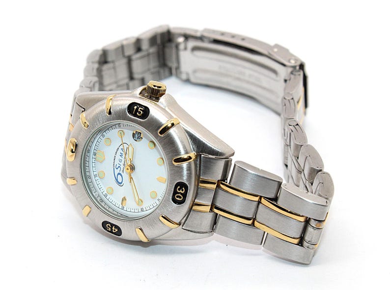 May include: A silver-toned wristwatch with gold accents. The watch face is white with black numerals and the brand name "Sigma". The watch band is silver with gold-toned links. The watch is angled to show the face and band.