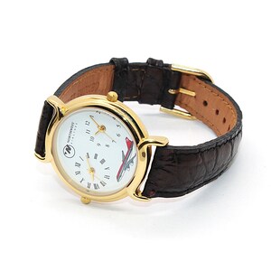 May include: A gold-toned wristwatch with a white face featuring the Northwest Airlines logo and a red airplane graphic. The watch has a brown leather band with a gold buckle. The watch is a vintage aviation collectible.