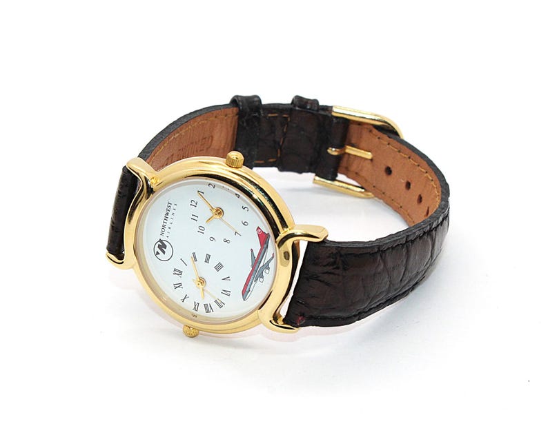 May include: A gold-toned wristwatch with a white face featuring the Northwest Airlines logo and a red airplane graphic. The watch has a brown leather band with a gold buckle. The watch is a vintage aviation collectible.