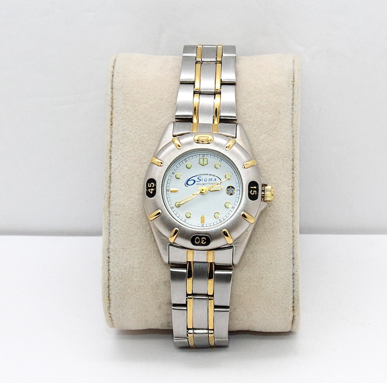 May include: A silver and gold-toned Sigma watch with a white face. The watch has a silver band with gold accents and a gold crown. The watch face displays the brand name and the numbers 15, 30, and 45. The watch is displayed on a beige cushion.