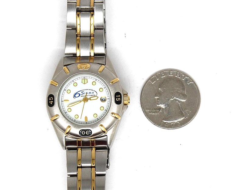 May include: A silver and gold-tone Sigma wristwatch with a white face, featuring black numerals and gold hands. The watch has a date display and is next to a US quarter coin.