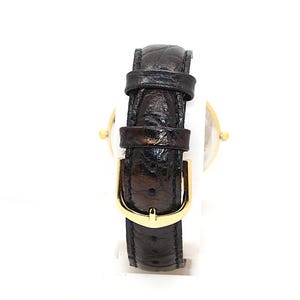 May include: A gold-toned wristwatch with a dark brown leather band. The band features a gold-colored buckle. The watch is set against a white background, highlighting the details of the watch.