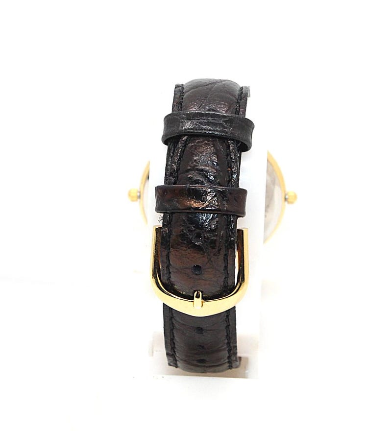 May include: A gold-toned wristwatch with a dark brown leather band. The band features a gold-colored buckle. The watch is set against a white background, highlighting the details of the watch.