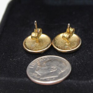 May include: A pair of gold-toned round stud earrings with a textured surface. The earrings are shown on a black velvet background next to a US dime.