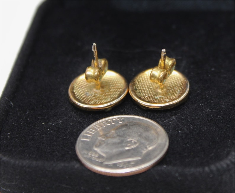 May include: A pair of gold-toned round stud earrings with a textured surface. The earrings are shown on a black velvet background next to a US dime.