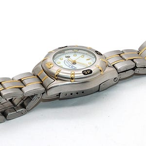 May include: A silver and gold-tone wristwatch with a white face. The watch has a round face with black markings and a silver and gold-tone band. The watch is lying on a white surface.