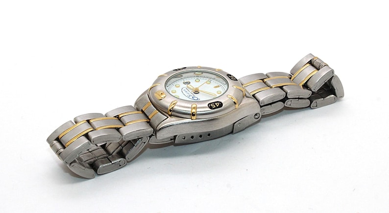 May include: A silver and gold-tone wristwatch with a white face. The watch has a round face with black markings and a silver and gold-tone band. The watch is lying on a white surface.