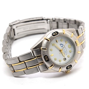 May include: A silver-toned wristwatch with gold-colored accents. The watch face is white with black numerals and the brand name "SIGMA". The watch band is a combination of silver and gold-colored links.