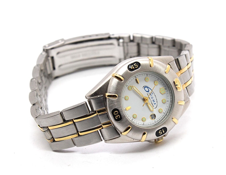 May include: A silver-toned wristwatch with gold-colored accents. The watch face is white with black numerals and the brand name "SIGMA". The watch band is a combination of silver and gold-colored links.