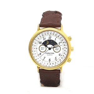 Op de afbeelding: Goudkleurig Carlton Quartz horloge met een witte wijzerplaat, met een maanfase-indicator en dag/datum-weergave. Het heeft een bruine leren band en drie knoppen aan de zijkant. De wijzerplaat toont de tekst "CARLTON QUARTZ".