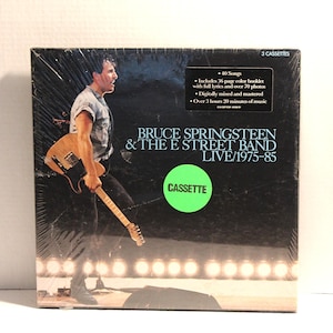 Puede incluir: Caja de casetes sellada de Bruce Springsteen & The E Street Band Live/1975-85. La portada presenta una foto de Springsteen tocando la guitarra en el escenario. El set de cajas incluye 3 casetes y un folleto a color de 36 páginas.
