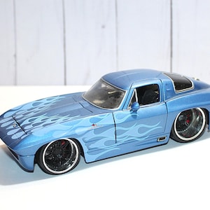 May include: A blue die-cast model car featuring a light blue flame pattern across the bonnet and sides. The vehicle has a low-profile stance, black wheels with silver rims, a sleek coupe body style, and clear windows revealing a dark interior.